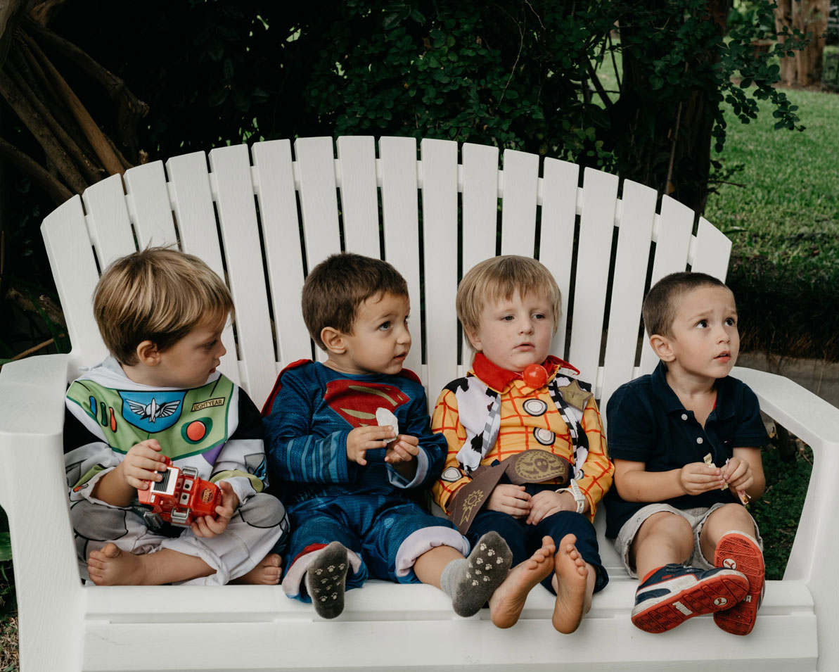 Four little boys in superhero and cowboy costumes sitting on a white garden bench at a birthday party in Haarlem