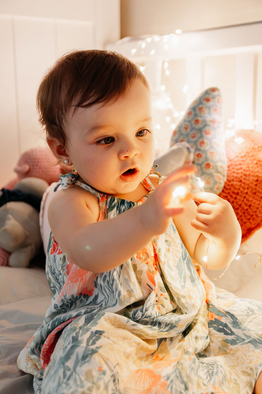 Baby girl in a floral dress reaching for twinkling fairy lights on her bed during a cozy winter mini session in Haarlem