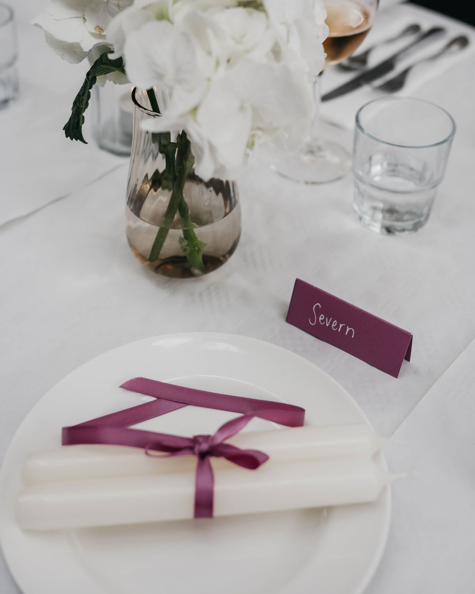 Place setting with a name card for Severn and white candles tied with a burgundy ribbon