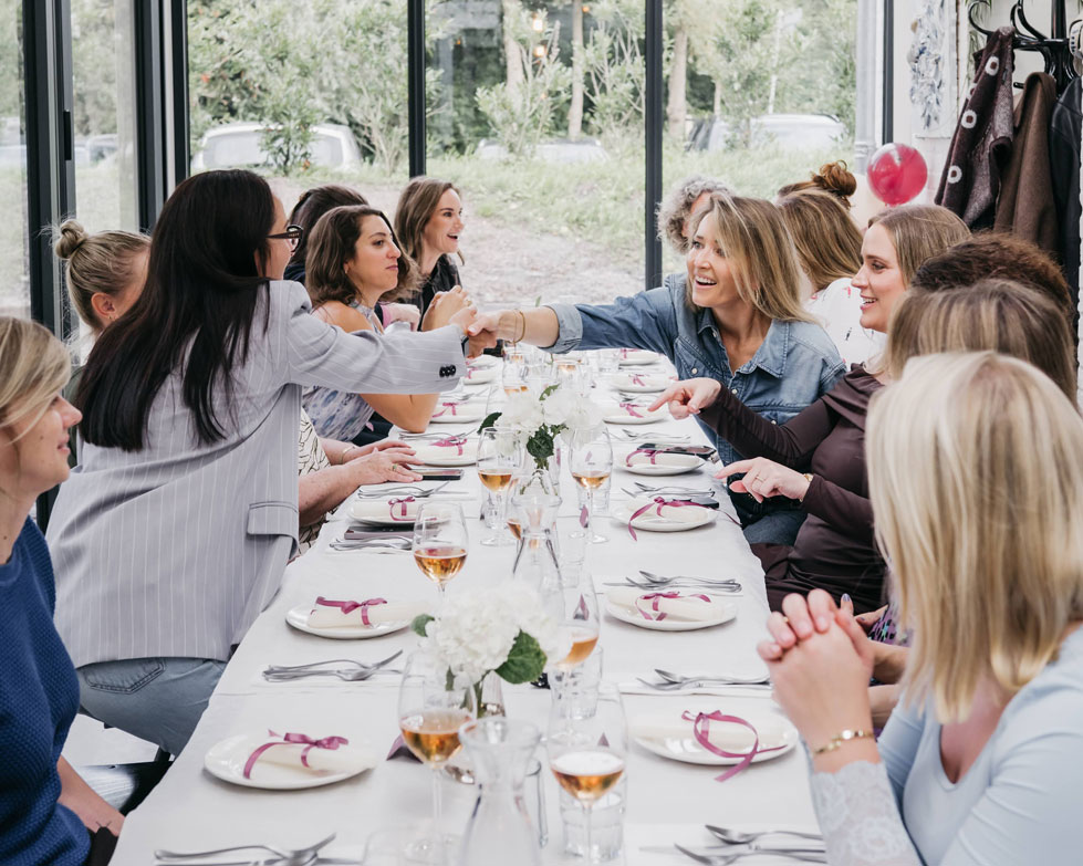 Women laughing and greeting each other around a long dinner table at a celebration lunch