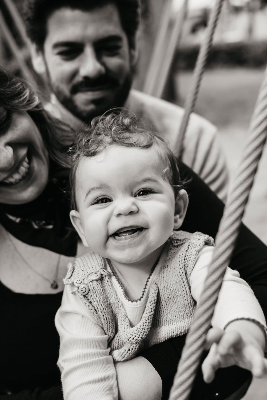 Baby girl laughing on a swing with her parents behind her in Westerpark, Amsterdam