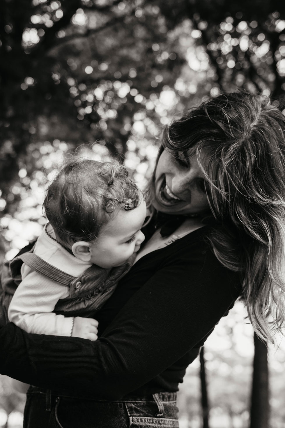 Mother laughing with her hair blowing in the wind while holding her baby under the trees